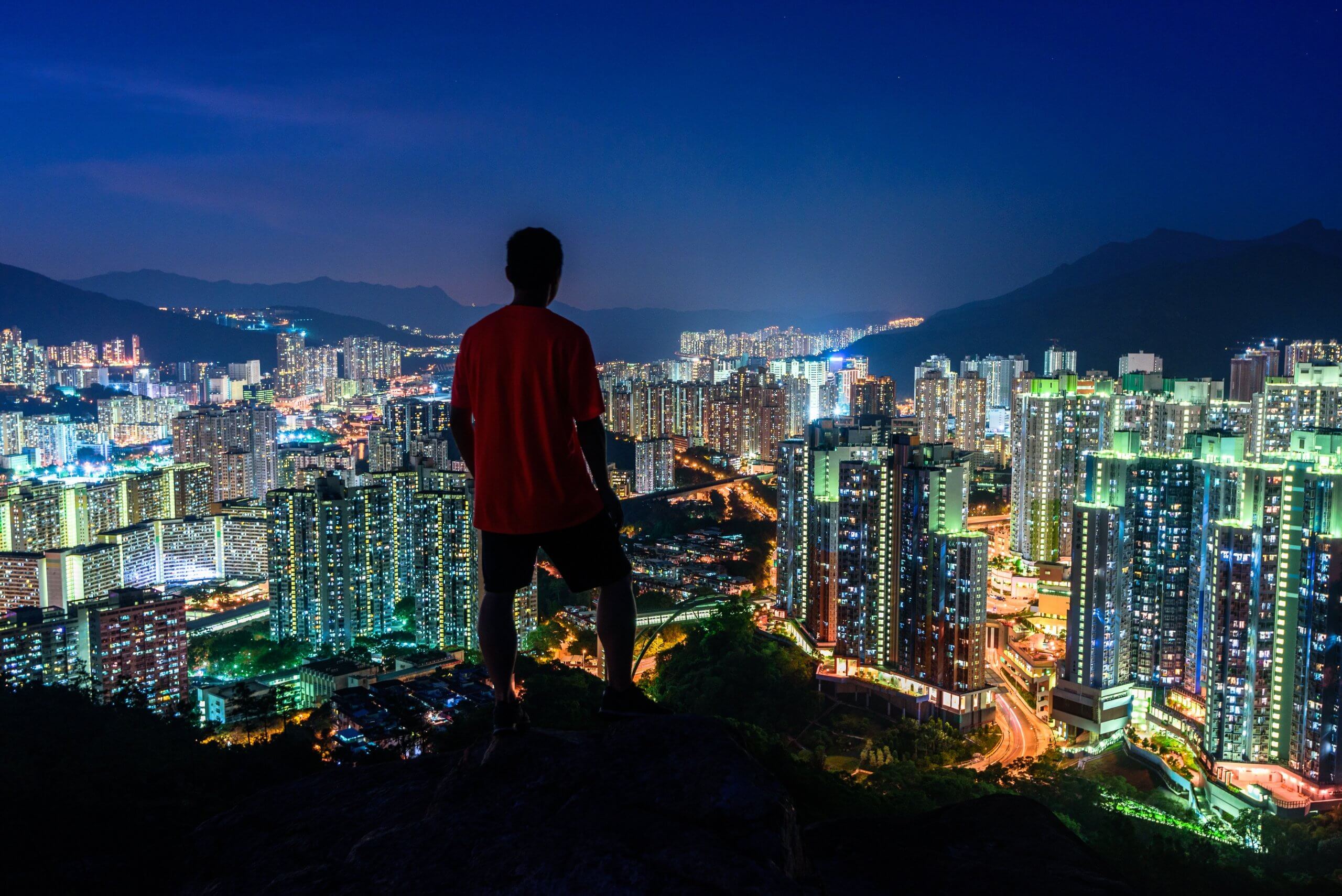 Person standing on a cliff looking over the lighted skyscrapers at night captured in Hong Kong