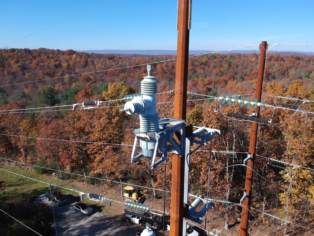 High-voltage power lines and insulators on a transmission tower in a forested area with autumn foliage.
